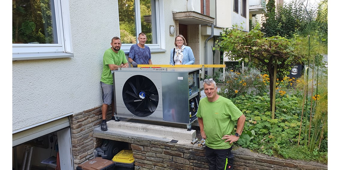 Eine Wärmepumpe wird rechtwinklig zu einem Mehrfamilienhaus auf einer Mauer zur Kellereinfahrt montiert, daneben stehen Mitarbeiter der Installationsfirm und die Bürgermeisterin