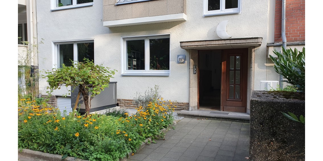 Eine Wärmepumpe steht im Vorgarten eines Mehrfamilienhauses rechtwinklig zur Außenwand. Im Vorgarten blühen gelbe Blumen und ein kleiner Baum steht vor der Wärmepumpe.