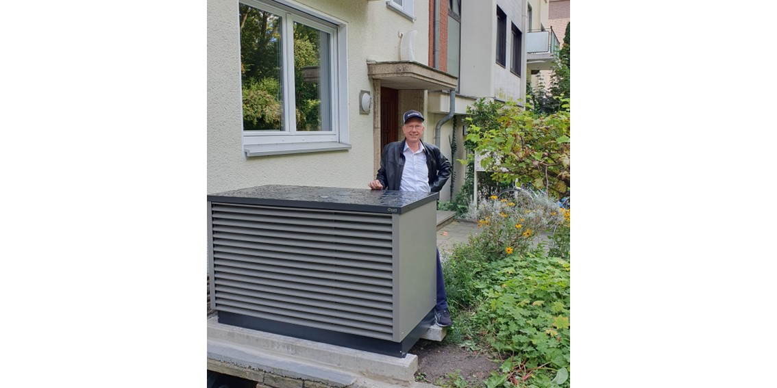 Eine Wärmepumpe steht im Vorgarten eines Mehrfamilienhauses, dahinter ein Mann mit schwarzer Jacke und hellem Hemd