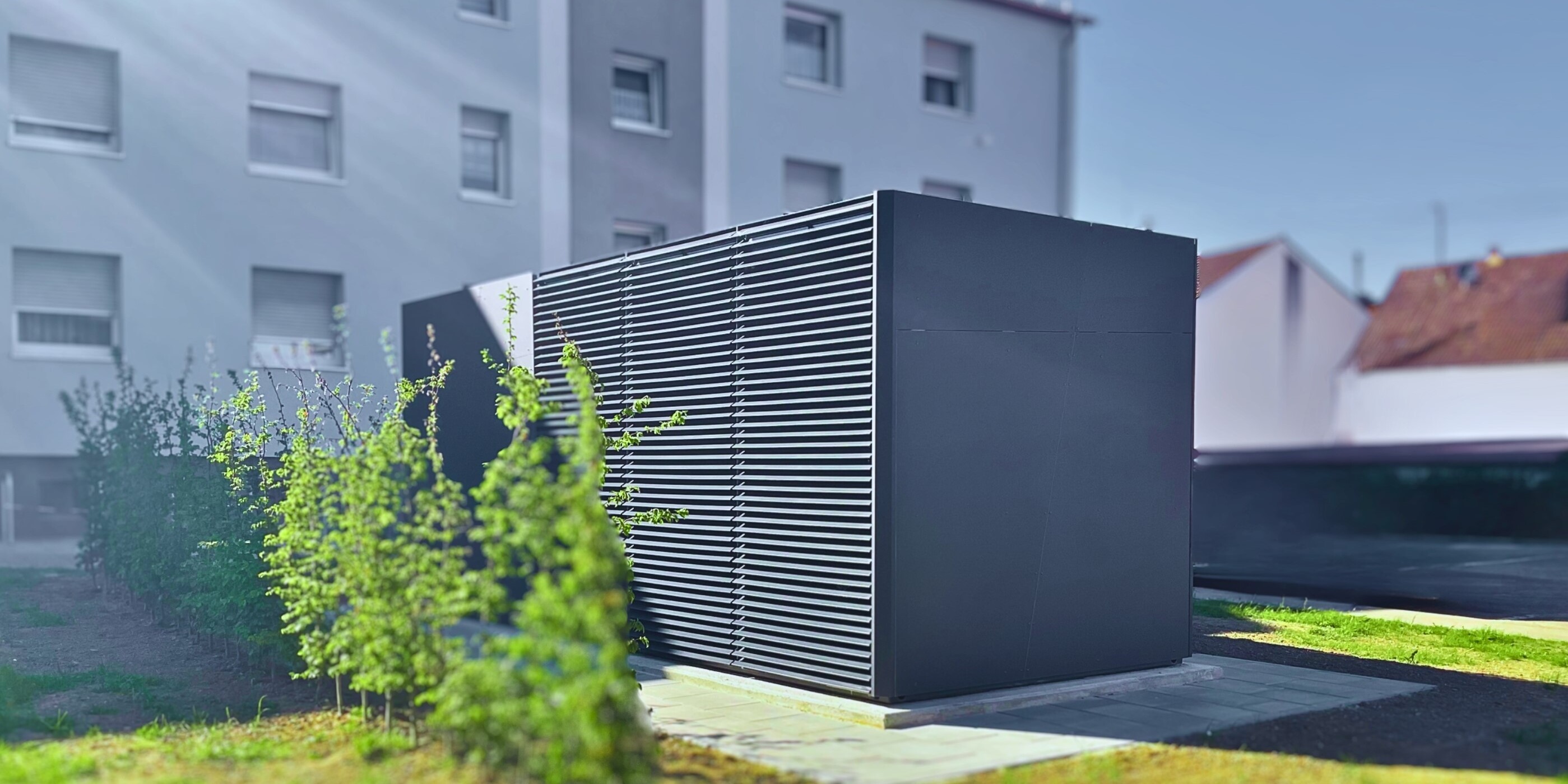 Die Wärmepumpenzentrale, ein dunkelgrauer Container mit Lüftungsschlitzen, ist von schräg vorne zu sehen, im Hintergrund teilweise das dreistöckige, hellgraue Mehrfamilienhaus.