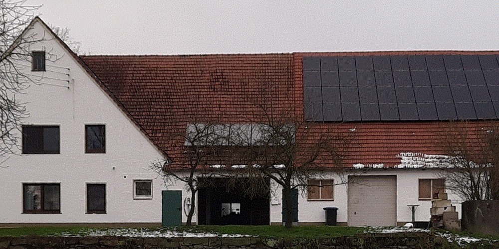 Ein großes Bauernhaus mit Solarkollektoren und Photovoltaik auf dem Dach vor teils verschneiter Wiese mit kahlen Obstbäumen