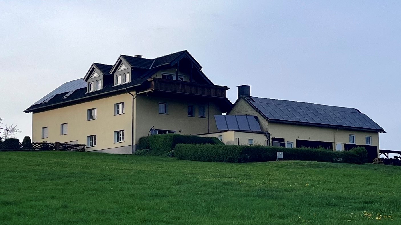 Außenansicht eines Gebäudes auf einer Wiese vor blauem Himmel, inkl Solarthermie und Photovoltaik auf dem Dach