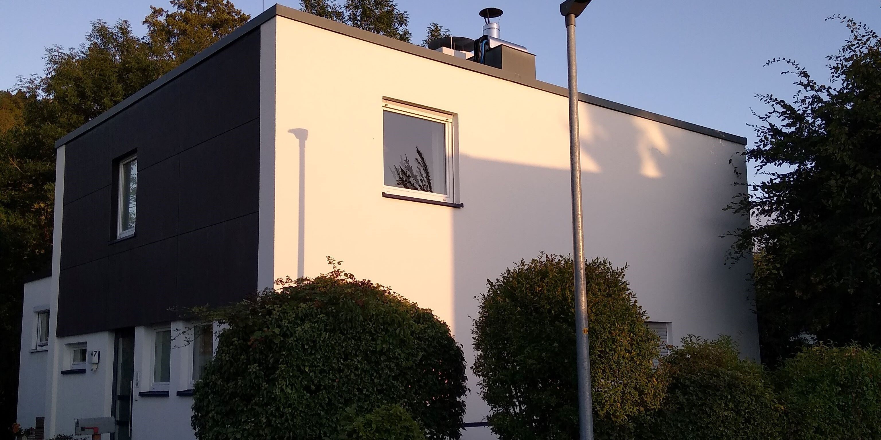 Ein zweigeschossiges, weißes Einfamilienhaus mit Flachdach, schräg von der Straße gesehen; das Obergeschoss über dem Eingang ist mit dunklem Holz verkleidet