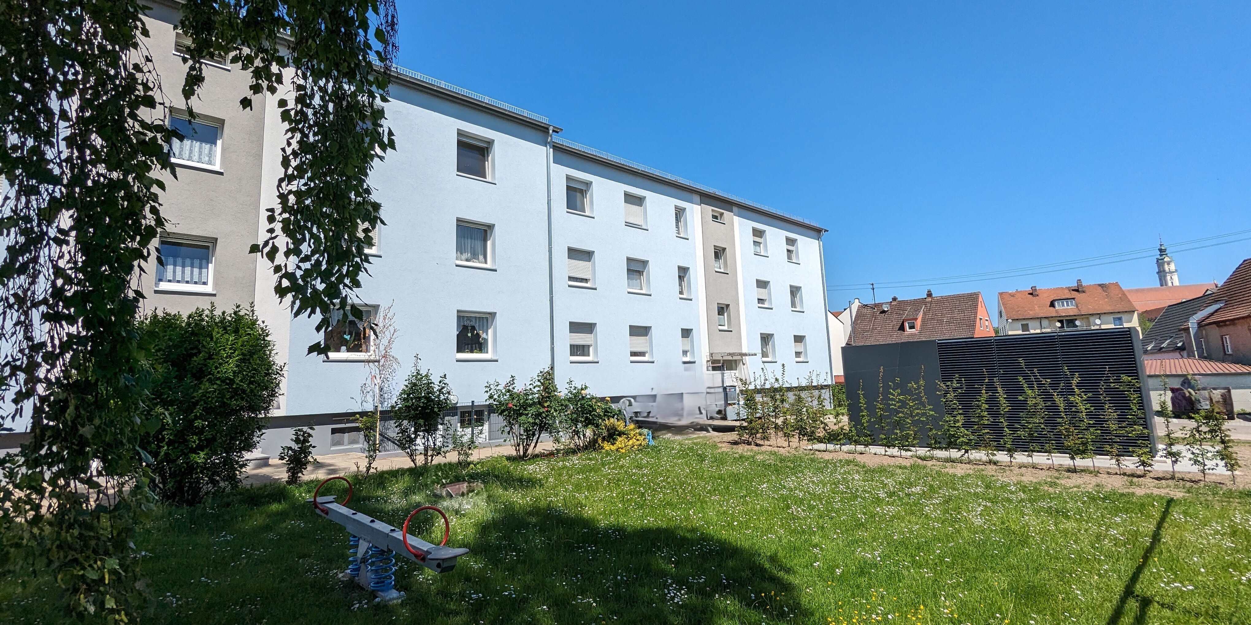 Ansicht fast des gesamten Innenhofes mit dem Container der Wärmepumpenzentrale auf der Wiese und links das dreistöckige, hellgraue Mehrfamilienhaus.