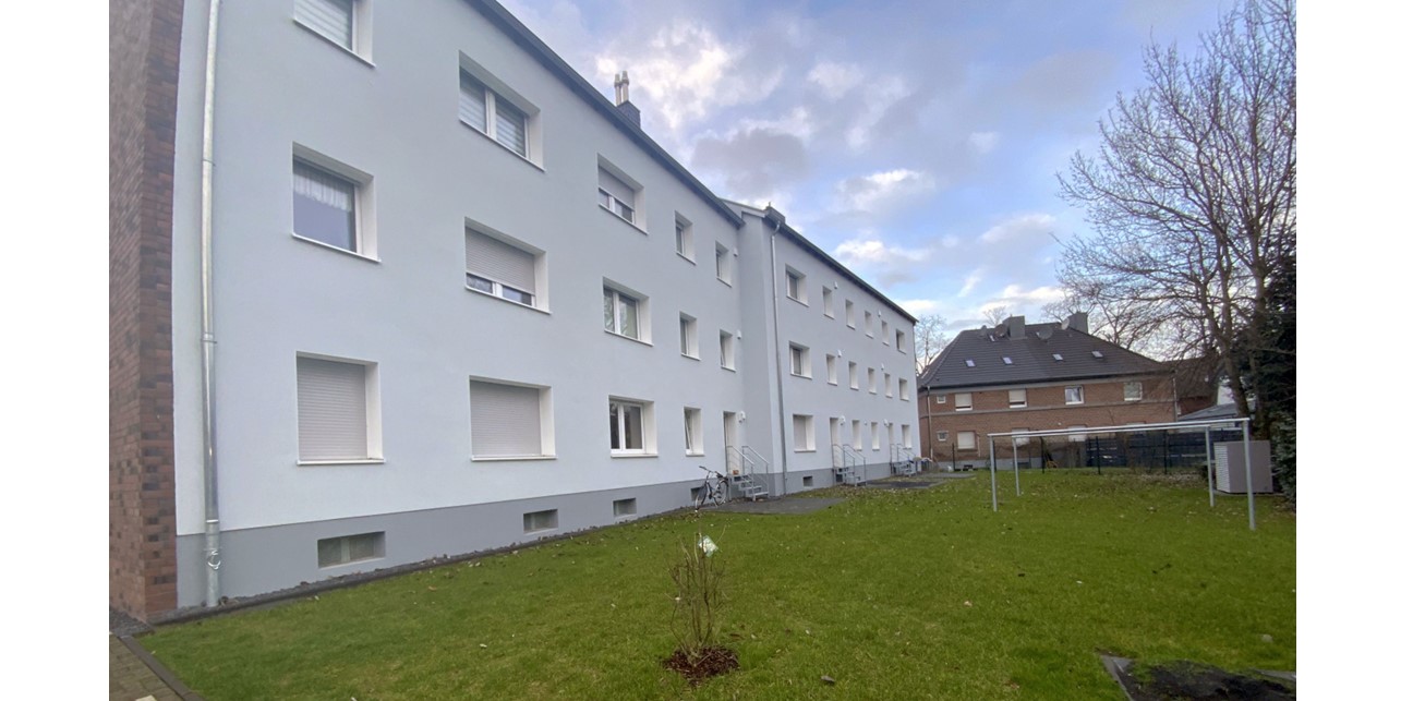 Ein dreistöckges, hellgraues Mehrfamilienhaus, schräg von hinten gesehen. Davor ist eine Wiese mit Wäscheplatz. Ein zweites ähnliches Haus schließt sich seitlich an.