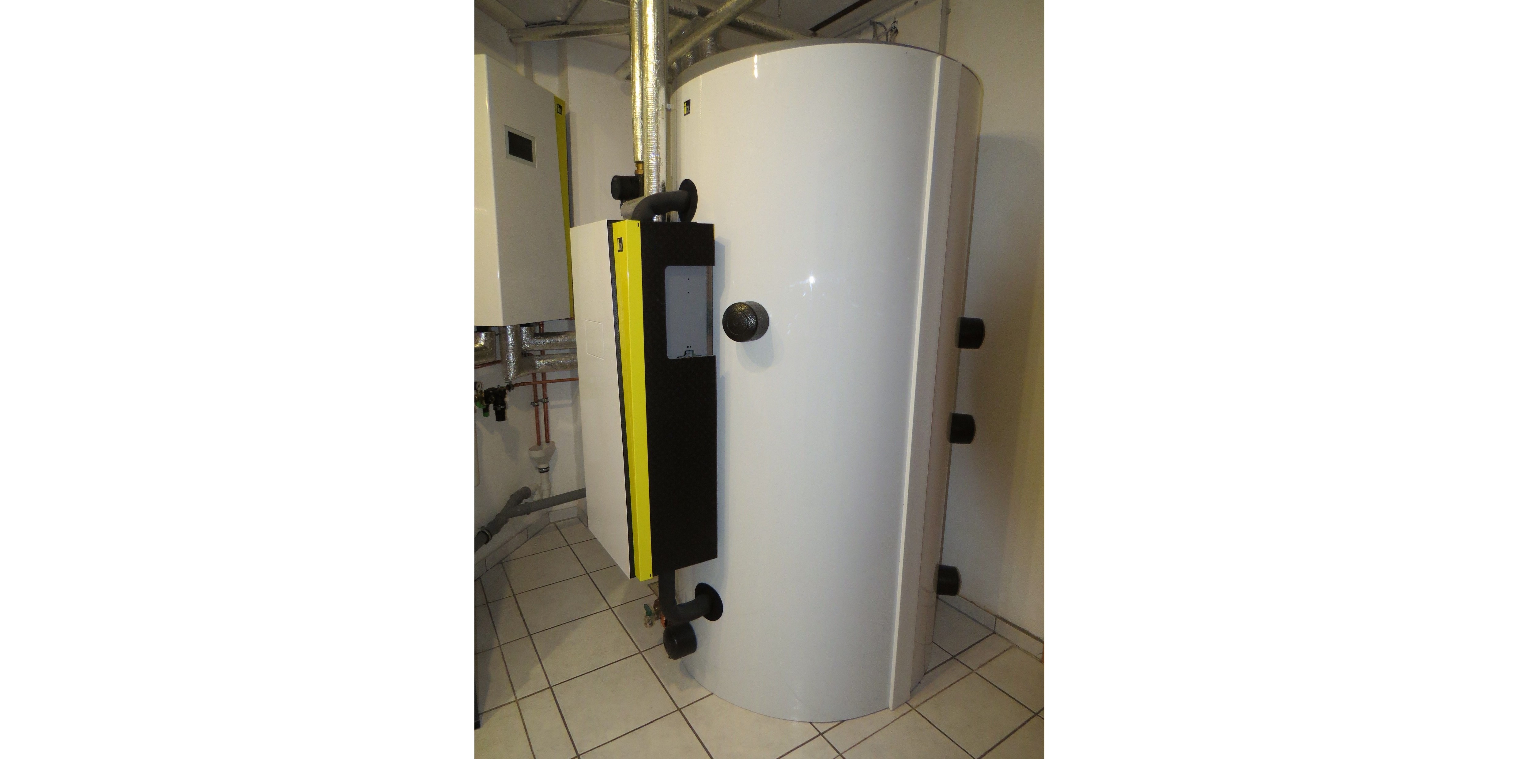Ein hellgrauer, fast raumhoher Pufferspeicher. Der Kasten mit dunkelgrauem und gelbem Streifen an der Vorderseite enthält die Frischwasserstation.
