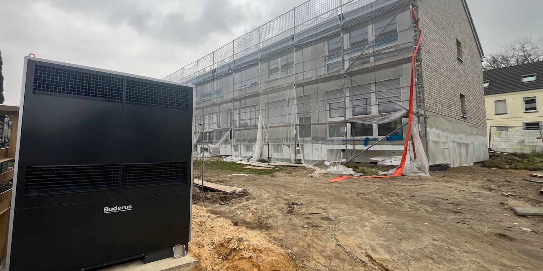 Schrägansicht des eingerüsteten Mehrfamilienhauses mit der Wärmepumpe vorne links im Vordergrund