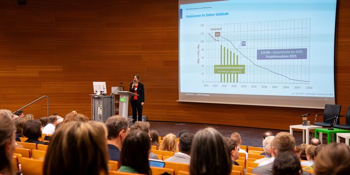 Carla Vollmer steht auf der Bühne im holzvertäfelten UBA-Hörsaal und spricht in ein Mikrofon. Im Vordergrund sind unscharf die Hinterköpfe des Publikums abgebildet. Im Hintergrund wird eine Präsentation projiziert.