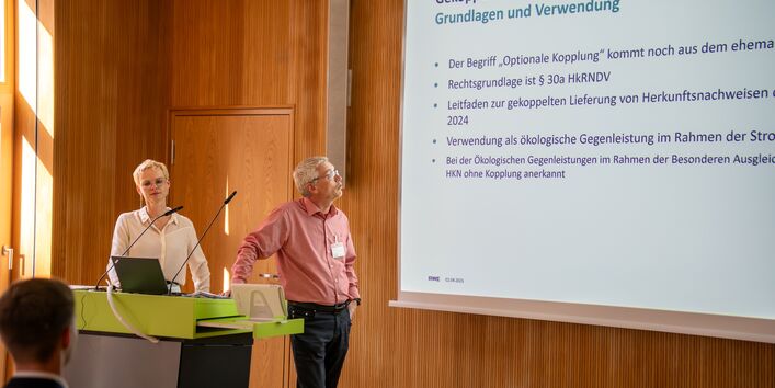 Eine Person referiert mit Blick auf eine projizierte Präsentation. Eine weitere Person steht daneben. Im Vordergrund ist unscharf ein Hinterkopf abgebildet.