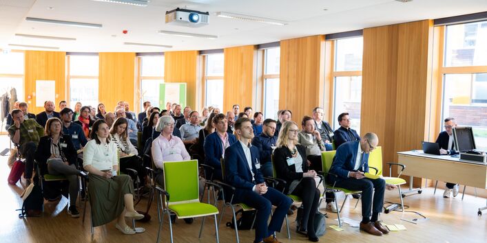 Etwa 40 Menschen sitzen auf Stühlen in einem Sitzungsraum und blicken interessiert nach vorn.