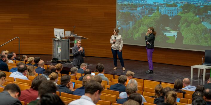 Eine Person spricht in der ersten Reihe des UBA-Hörsaals zum Publikum gewandt in ein Mikrofon. Zwei weitere Personen stehen zuhörend auf der Bühne. Im Vordergrund sind unscharf die Hinterköpfe des Publikums abgebildet. Im Hintergrund wird eine Präsentation projiziert.