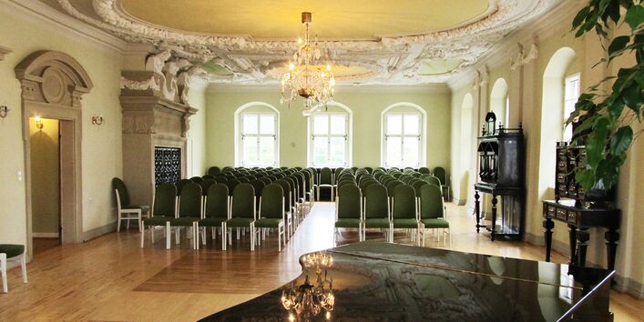 Barocksaal mit Stuckdecke und Holzparkettboden, im Vordergrund steht ein schwarzer Flügel, der Raum ist bestuhlt