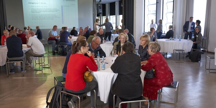 In einem großen Raum stehen mehrere runde Tischen, an denen diskutierende Menschen sitzen.