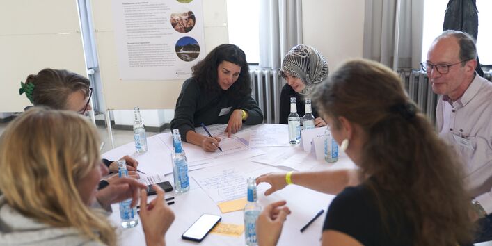 Das Foto zeigt einen Tisch mit mehreren Menschen, von denen eine Teilnehmerin Notizen macht.