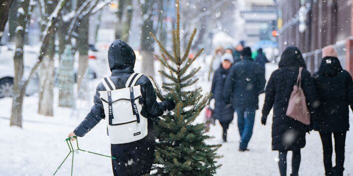 Ein Mensch läuft in einer verschneiten Stadt mit einem Weihnachtsbaum in der Hand in der Fußgängerzone entlang.