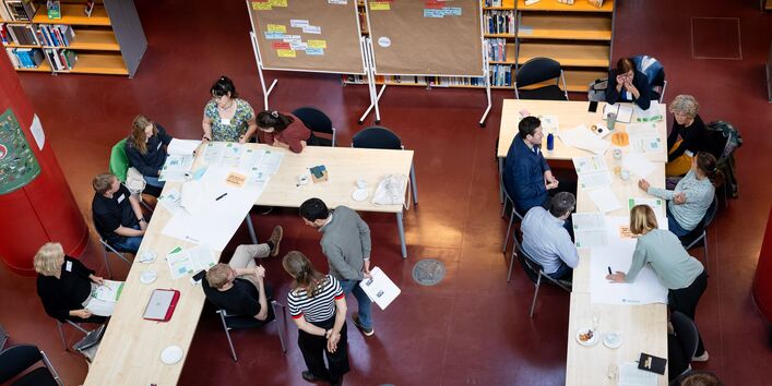 Das Foto zeigt Arbeitsgruppen mit Menschen an Gruppentischen, die sich austauschen und Inhalte schriftlich festhalten.
