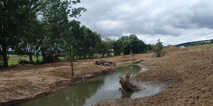 Foto: Die Wern mit deutlichen Spuren der kurz zuvor beendeten Baumaßnahmen. Die Ufer bestehen noch aus offenem Boden und sind frei von Vegetation. Im Gewässer liegt ein in die Sohle eingebauter Wurzelstock.