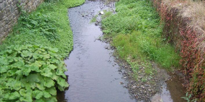 Foto: Bach mit hohen Ufermauern und angrenzenden Häusern und Straßen in Ortslage. Die Gewässersohle ist offen, mit Kies bedeckt und stellenweise bewachsen. Der Bach verläuft leicht schwingend im Kastenprofil.