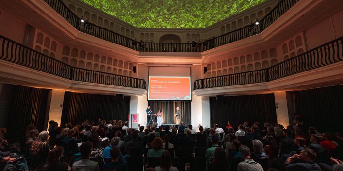 Der Hauptraum des ehemaligen Krematoriums ist voll besetzt mit TeilnehmerInnen. Auf einer Bühne findet das Eröffnungsgespräch statt.