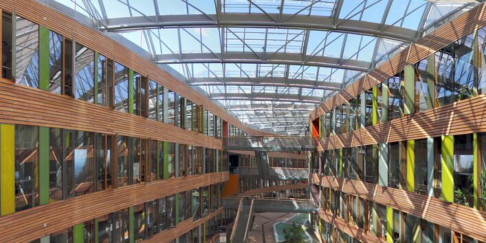 Blick in den mit Glas überdachten Innenhof, rechts und links die Innenfassaden mit Holzvertäfelungen und Glasscheiben in Grüntönen, im Innenhof Brücken und Treppen, die die Gebäudeteile verbinden