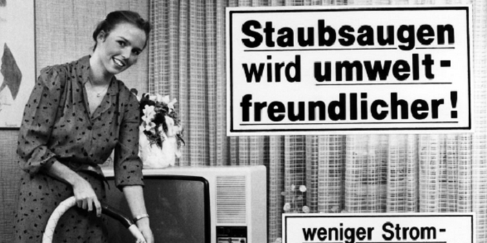 Das alte Schwarz-weiß-Foto zeigt eine Frau beim Staubsaugen im Wohnzimmer. Aufschriften: Staubsaugen wird umweltfreundlicher, weniger Strom bei gleicher Leistung! weniger Lärm (max. 73 dB(A). Der Staubsauger trägt das Umweltzeichen Blauer Engel.