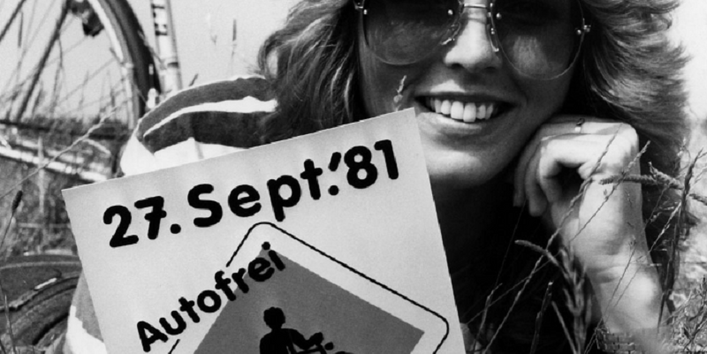 eine Frau lmit Sonnenbrille iegt neben ihrem Fahrrad auf einer Wiese und hält ein Schild in die Kamera: 27. Sept.´81, Autofrei – Spaß dabei“