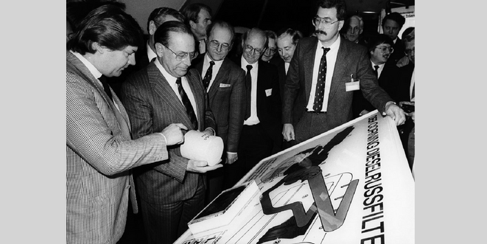 Männer in Anzügen stehen dicht gedrängt vor einer Schautafel zum Dieselrußfilter