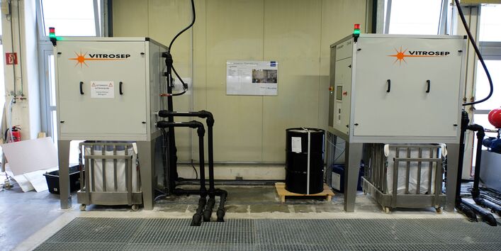 teschnische Anlage in einer Werkshalle aus zwei Boxen, aus denen Rohre in eine Gitterabdeckung im Fußboden führen