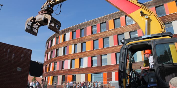 UBA-Präsident Flasbarth sitzt vor dem Dienstgebäude in Dessau-Roßlau in einem Bagger, um eine alte Wand aus Ziegeln einzureißen, Menschen am Bauzaun schauen zu