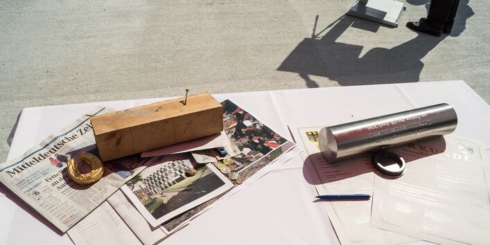 auf einem Tisch liegen eine Metallkapsel, eine Tageszeitung, Geldmünzen, Urkunden, Bauunterlagen und weitere Dokumente