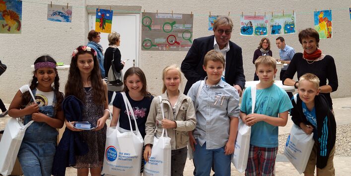 vier Mädchen und drei Jungen stehen in einer Reihe lächelnd vor der Kamera mit gefüllten Baumwollbeuteln mit der Aufschrift "Die BMU Klimaschutzinitiative". Dahinter Dr. Thomas Holzmann und Maria Krautzberger und die Kinderbilder auf der Wäscheleine.