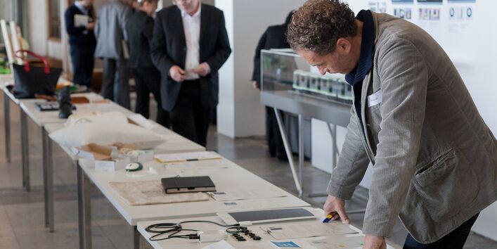 Auif einer langen Tischreihe liegen Produkte und Ausdrucke, die von mehreren Personen angeschaut werden