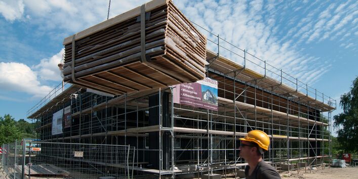 eingerüstetes Haus, davor ein Bauarbeiter mit gelbem Helm, ein Kran liefert Elemente für die Holz-Fassadenverkleidung an