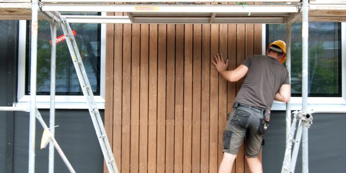 zwei Bauarbeiter stehen auf einem Baugerüst und montieren zusammen ein Holz-Fassadenelement