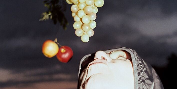 von oben hängen Weintrauben und Äpfel herab, darunter eine junge Frau mit Kapuzenjacke, die versucht, mit dem Mund heranzukommen, im Hintergrund Dämmerung und dunkle Wolken