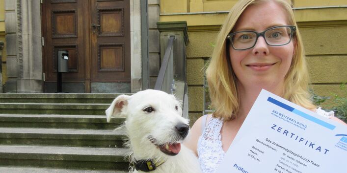 eine Frau hält mit der einen Hand einen weißen Terrier und mit der anderen ein Blatt Papier mit der Aufschrift "Zertifikat"