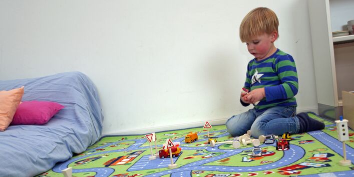 Kind spielt auf einem Teppich, der wie eine Straßenlandschaft aussieht, mit Spielzeugautos.