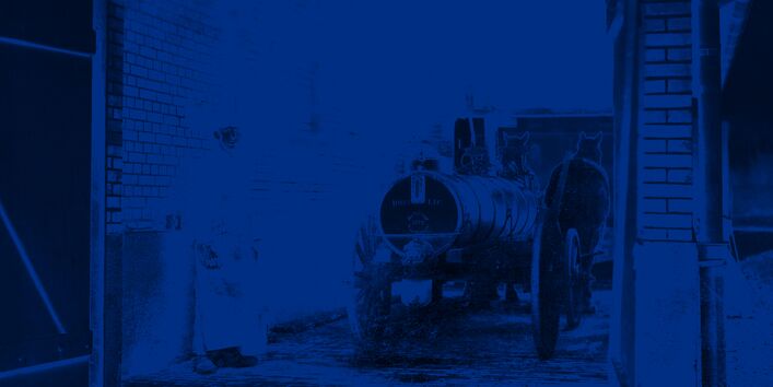 Ein Mann steht mit einem Jauchewagen in einer Einfahrt, der Wagen trägt einen Metalltank und wird von zwei Pferden gezogen. 