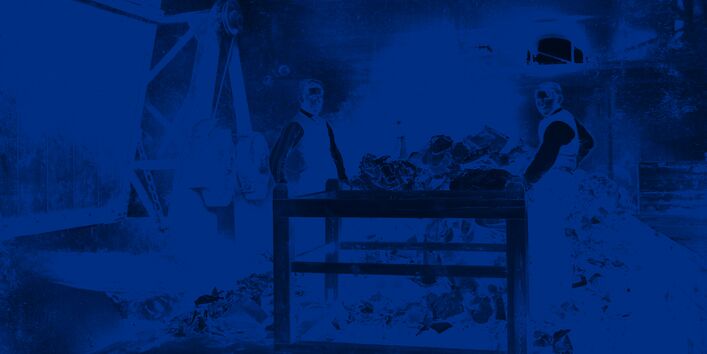 Zwei Arbeiter stehen zwischen einem Tisch, einem Berg Müll und einem Kran.