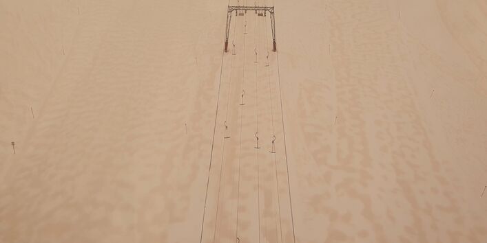 Auf dem Foto ist ein Schlepplift und Skipisten von oben zu erkennen. Auf dem Schnee sieht man großflächig bräunliche Sandablagerungen.