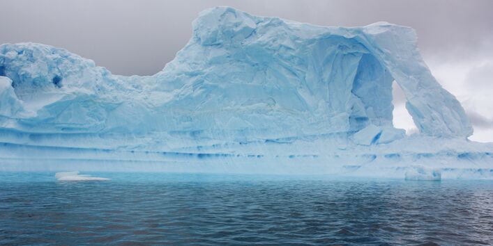 Im fast schwarzen Wasser schwimmt ein hellblau anmutender Eisberg. Der Himmel wirkt grau und trist.