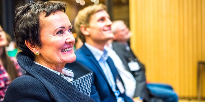 Es ist das Profil einer Frau zu sehen. Sie schaut nach vorne und lacht. Sitzt mit anderen Leuten in einer Stuhlreihe im Publikum und auch der Mann links neben ihr grinst interessiert.