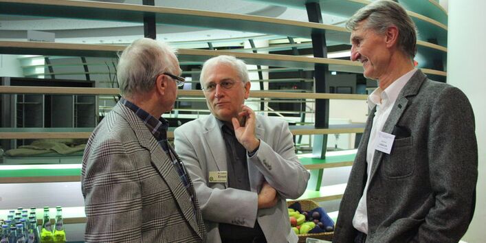 Drei  ältere Männer im Jacket stehen in einem Kreis zusammen und reden.