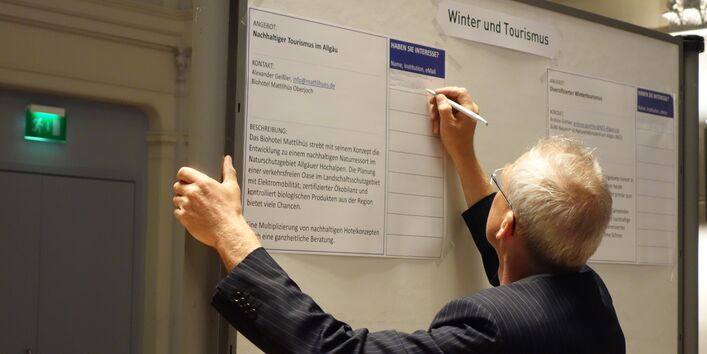 Ein Teilnehmer steht vor einer Informationstafel und hat einen Stift in der Hand. Er ist der Kamera mit dem Rücken zu gedreht und füllt eine vorgedruckte Tabelle auf der Informationstafel aus.