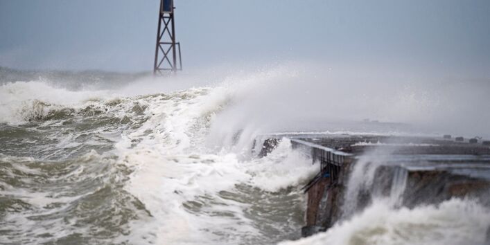 The picture shows a storm surge wave breaking over a coastal structure. A metal mast is visible in the background. 