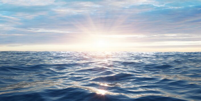 The picture shows a slightly agitated sea surface under a low sun. The sky is covered in bluish-grey clouds. 