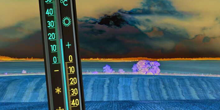 In the foreground the picture shows a thermometer indicating 20°C. The landscape in the background is characterised by intensive agriculture; a cereal field after harvesting is visible. Dark clouds cover the sun which lights up the sky in the background behind the dark clouds. 