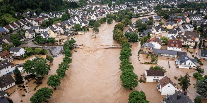 Das Bild zeigt eine Siedlung mit Einfamilienhäusern und kleinen Mehrfamilienhäusern entlang eines Flusstals. Der durch die Ortschaft fließende Fluss ist über die Ufer getreten und überschwemmt die tiefer liegenden Teile der Ortschaft mit bräunlichem Wasser. Der ursprüngliche Flusslauf ist anhand von uferbegleitenden Baumreihen zu erahnen. In der Mitte des Bildes ist eine zerstörte Brücke, die den Fluss querte, sichtbar.