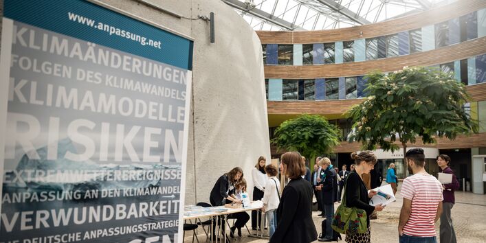 Blick ins Forum des Umweltbundesamt auf den Anmeldetisch. Personen befinden sich hinter und vor dem Anmeldebereich. Im Vordergrund steht ein Roll up beschriftet mit den Umweltrisiken und Chancen.