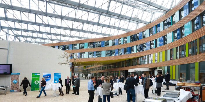 Veranstaltungsambiente im Forum des Umweltbundesamtes unter dem großzügigen Glasdach und vor der mit Holz verkleideten Fassade mit viel Fensterglas in den Farben Grün und Blau abgesetzt. Auf der Steinfläche am Boden stehen die Teilnehmenden an Stehtischen und diskutieren.